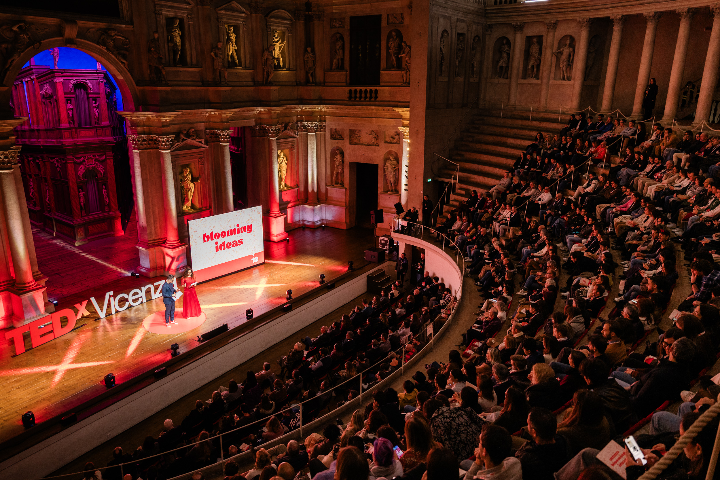 Pubblico di TEDxVicenza riunito in un teatro storico durante un evento, con relatori sul palco illuminato da luci rosse e una scenografia architettonica ricca di colonne e statue.
