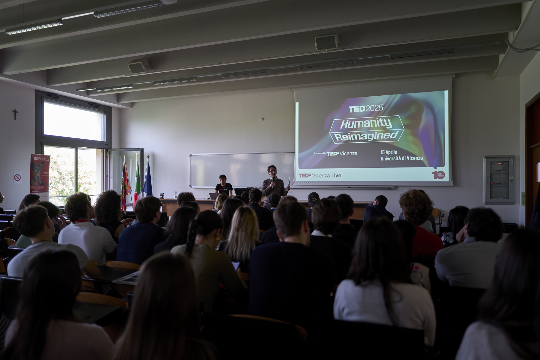 Pubblico riunito in un’aula dell’Università di Vicenza durante TEDxVicenza Live 2025, con due relatori davanti allo schermo che presenta il tema “Humanity Reimagined” del TED2025.