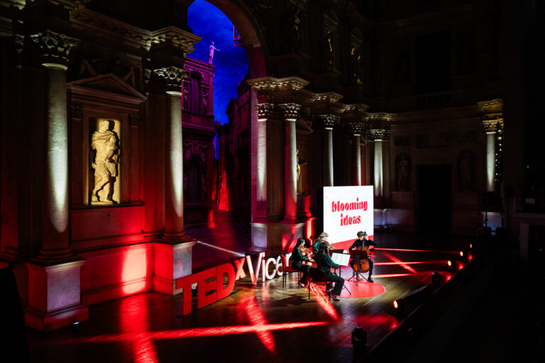 Quartetto d’archi si esibisce sul palco del TEDxVicenza Main Event 2025, allestito in un teatro storico con colonne e statue illuminate da luci rosse e viola e la scritta “Blooming Ideas” sullo schermo.