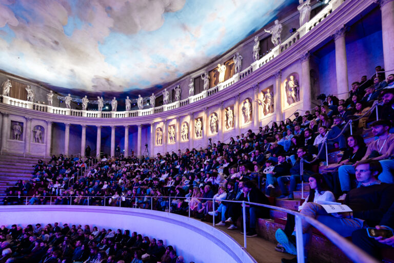 Pubblico numeroso del TEDxVicenza Main Event 2025 seduto sulle gradinate di un teatro storico, illuminato da luci blu e viola, durante l’evento.