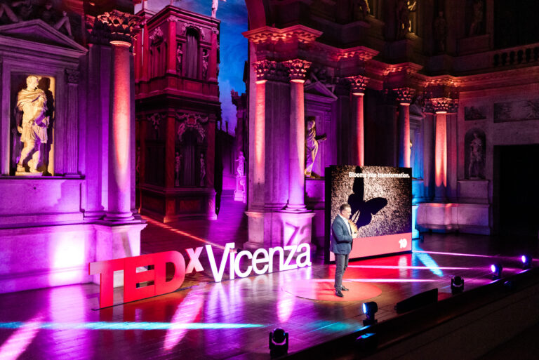 Relatore sul palco del TEDxVicenza Main Event 2025 durante un intervento sul tema “Blooming Ideas”, in un teatro storico illuminato con luci colorate e scenografia TEDxVicenza.
