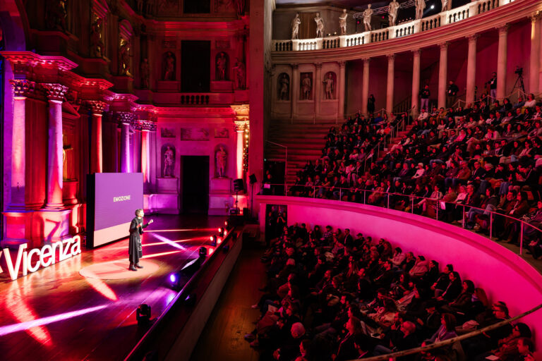 Relatrice sul palco del TEDxVicenza Main Event 2025 durante un intervento, davanti a un pubblico numeroso in un teatro storico illuminato con luci rosse e rosa.