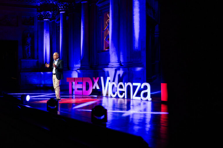 Relatore sul palco del TEDxVicenza Main Event 2025 durante un intervento, accanto alla grande scritta TEDxVicenza e circondato da colonne illuminate con luci blu e viola.