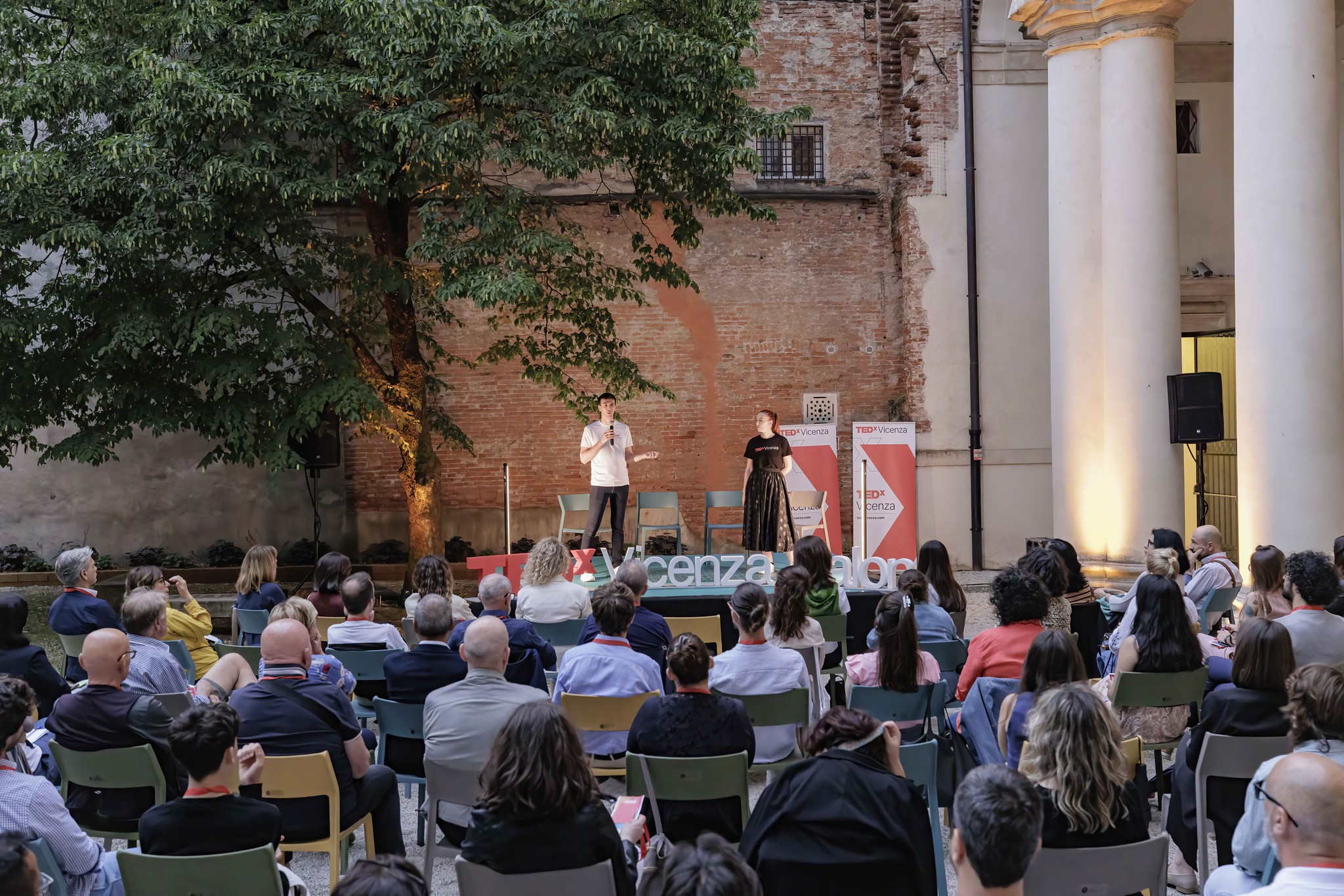 Pubblico riunito nel cortile storico di un edificio durante un TEDxVicenza Salon 2025, con due relatori sul palco e il pubblico seduto di fronte alla scenografia TEDxVicenza.